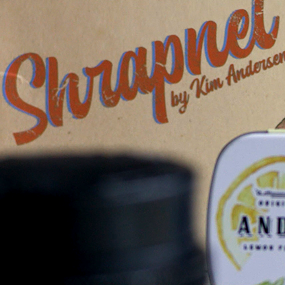The image shows the product packaging for 'Shrapnel' by Kim Andersen. The packaging is primarily black and orange with a illustration of a man wearing glasses and a beard.