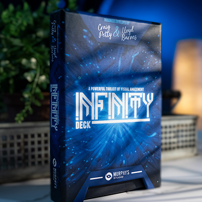 The image shows a boxed product labeled 'Infinity Deck' by Craig Petty and Lloyd Barnes. The box is open, revealing the blue interior with card designs, and is placed on a table with a plant and a decorative lamp in the background.