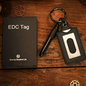 EDC Tag keychain on a wooden surface with a book and small box.